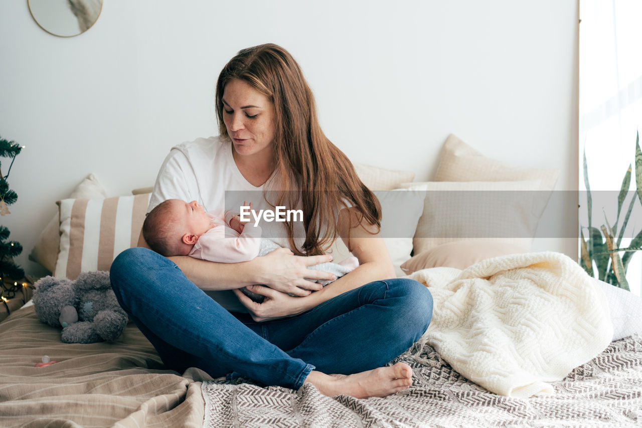 A tender caring mother lulls a newborn baby in her arms, sitting on the bed.