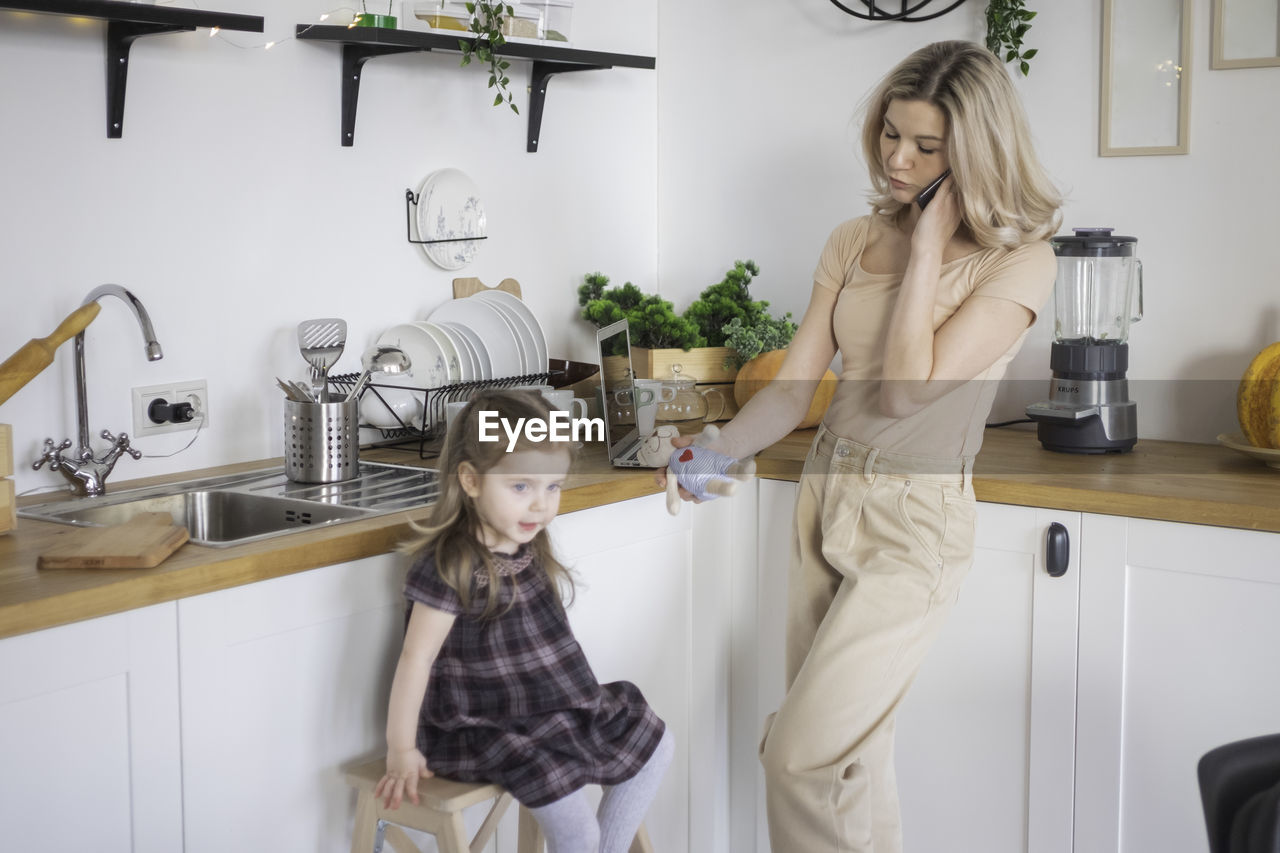 Mother talking on phone while daughter sitting stool in kitchen