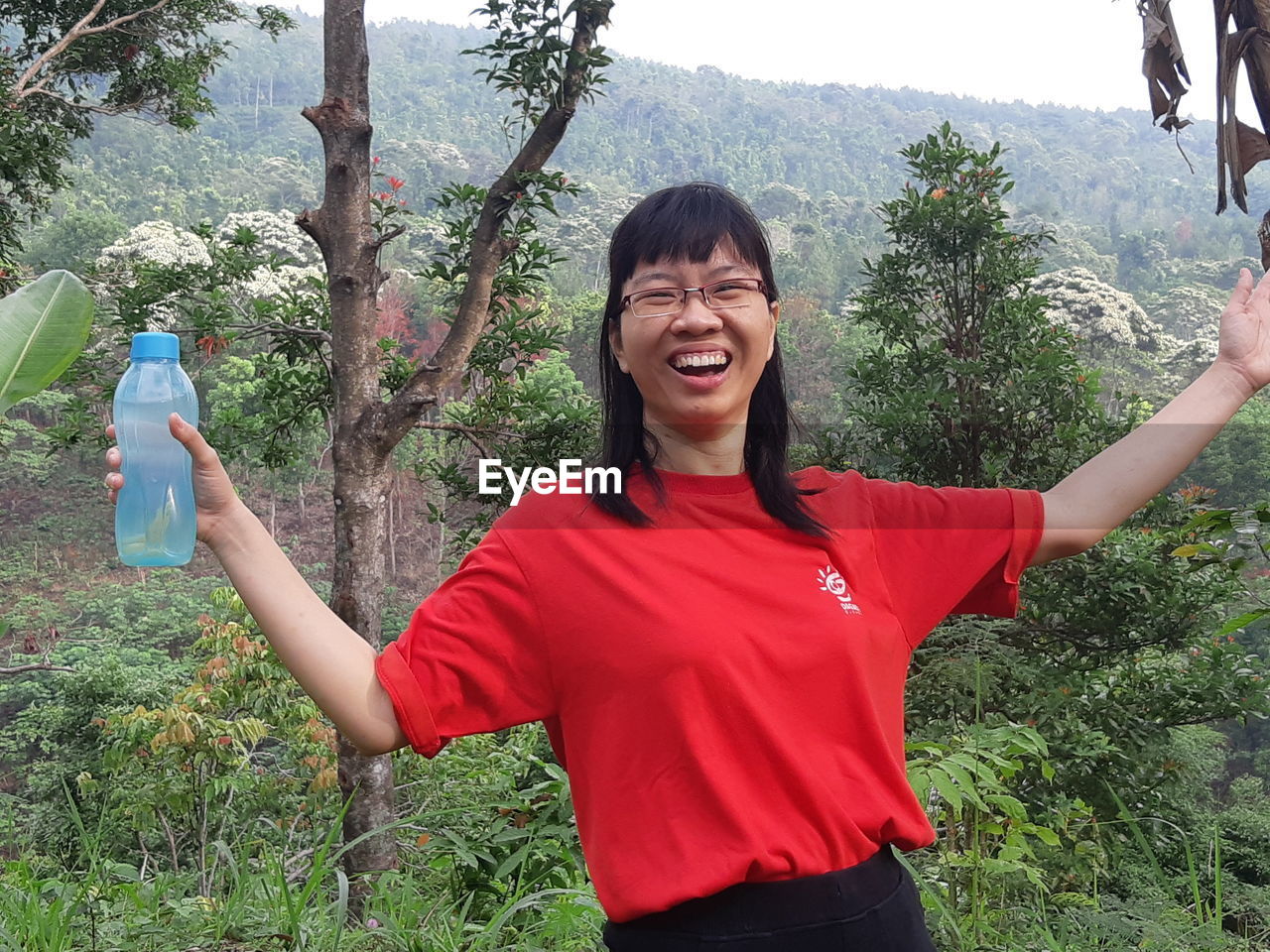 SMILING YOUNG WOMAN STANDING AGAINST PLANTS