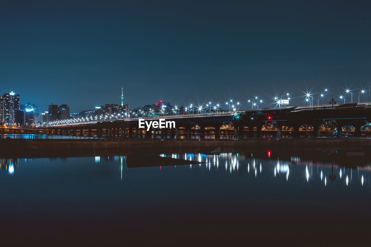 Illuminated bridge over river by buildings against sky at night