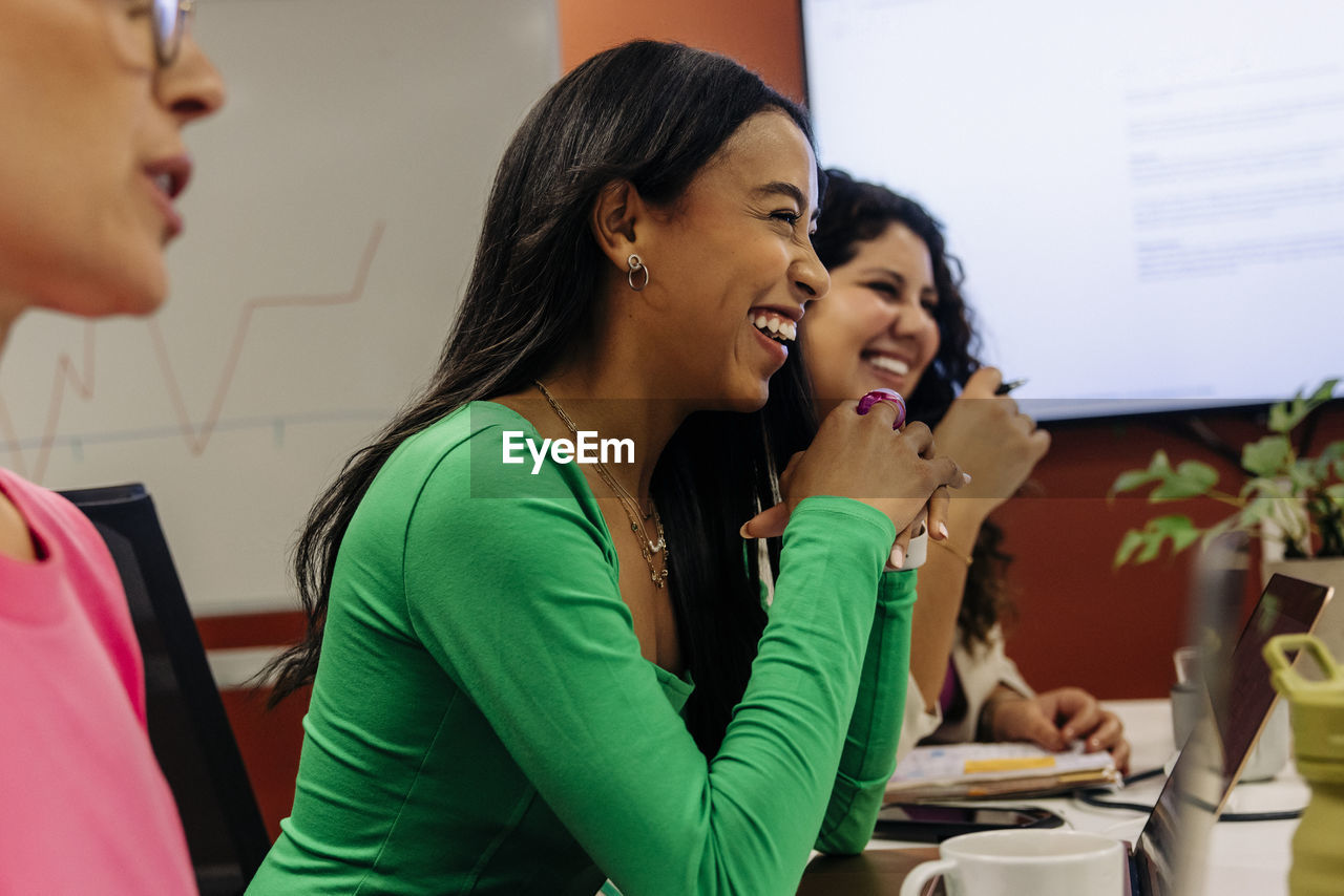 Happy female business professional leaning on elbows while sitting with colleagues during meeting at office