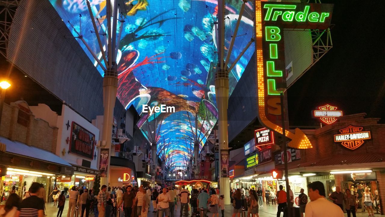 PEOPLE ON STREET AMIDST ILLUMINATED BUILDINGS IN CITY