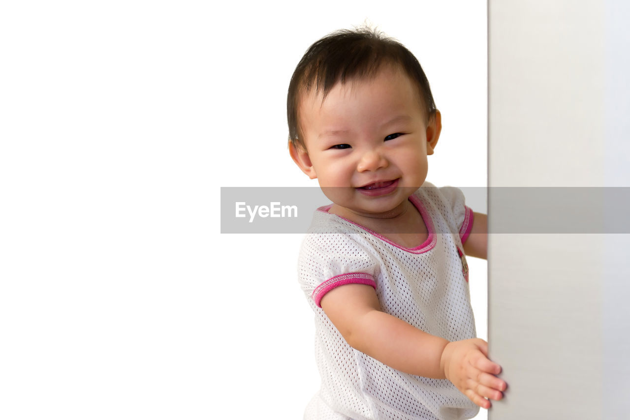 Close-up portrait of cute baby girl standing by wall against white background