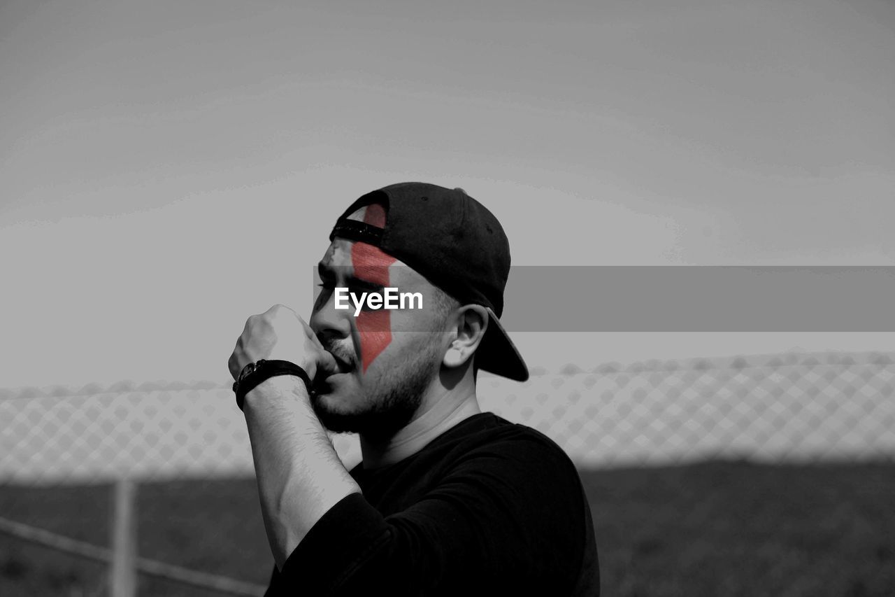 Thoughtful man with red face paint wearing cap against clear sky