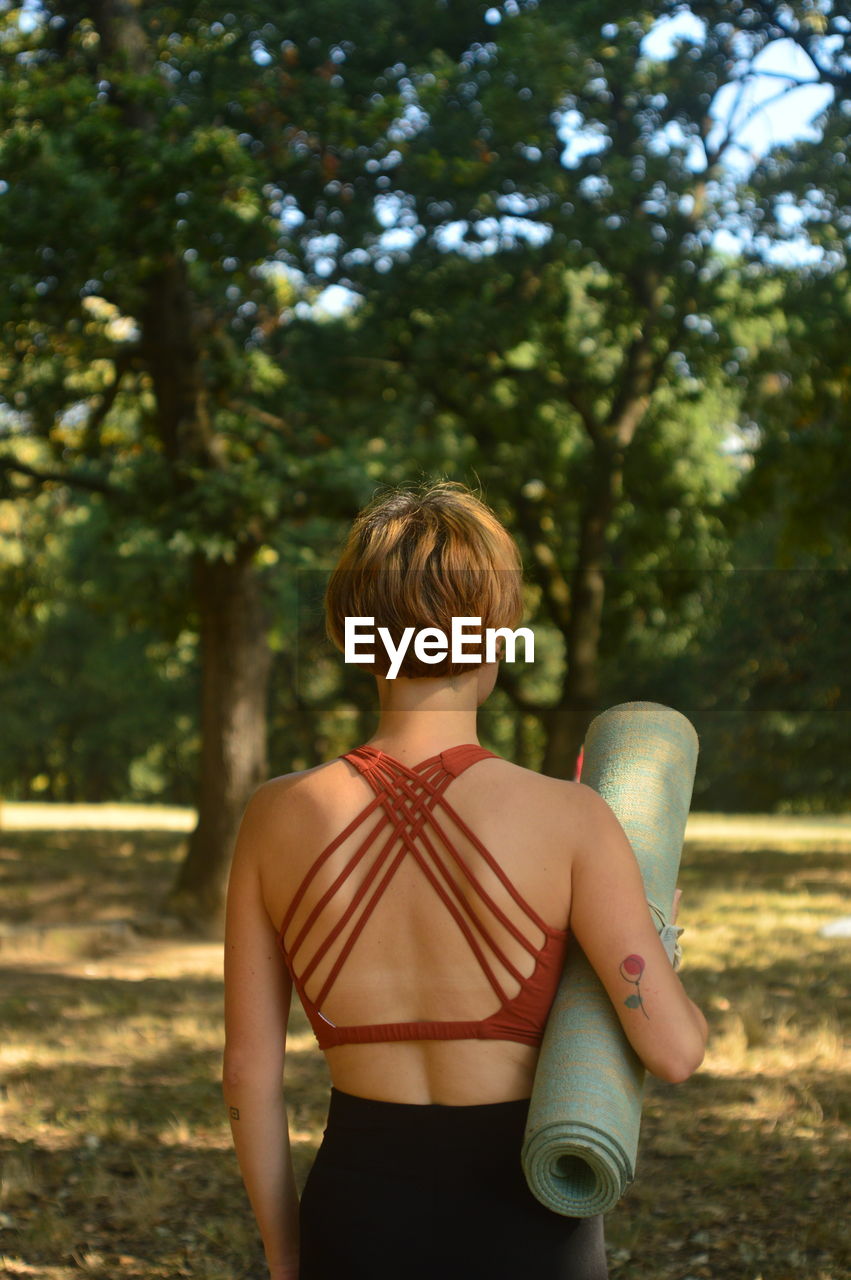 Rear view of woman standing with exercise mat against trees