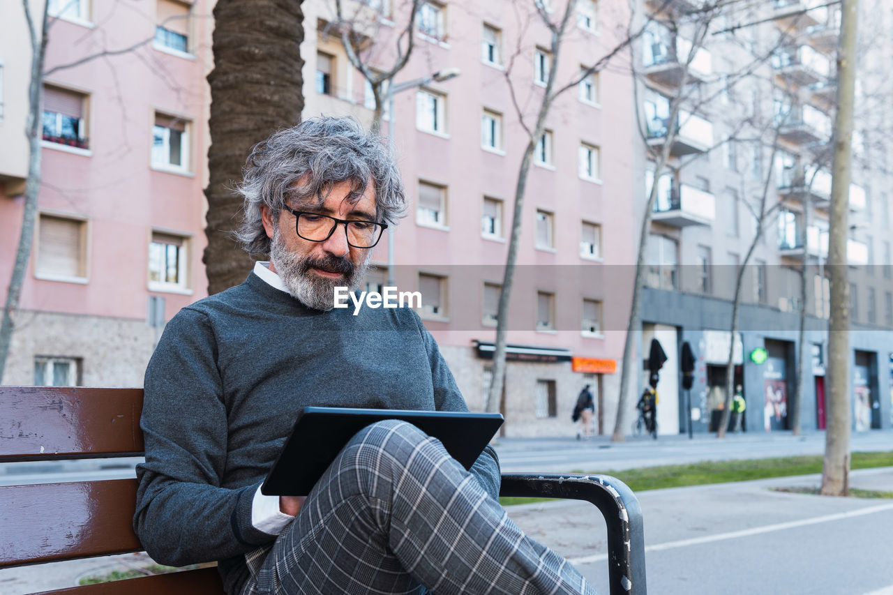 Handsome mature man in eyeglasses using digital tablet on bench in the street