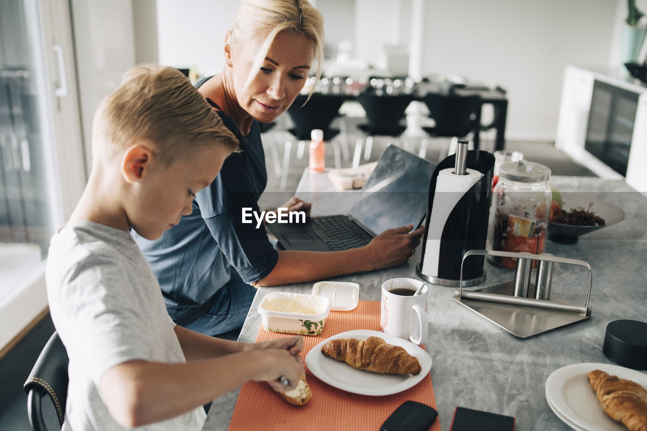 Mature woman with laptop looking at son applying butter on croissant in kitchen