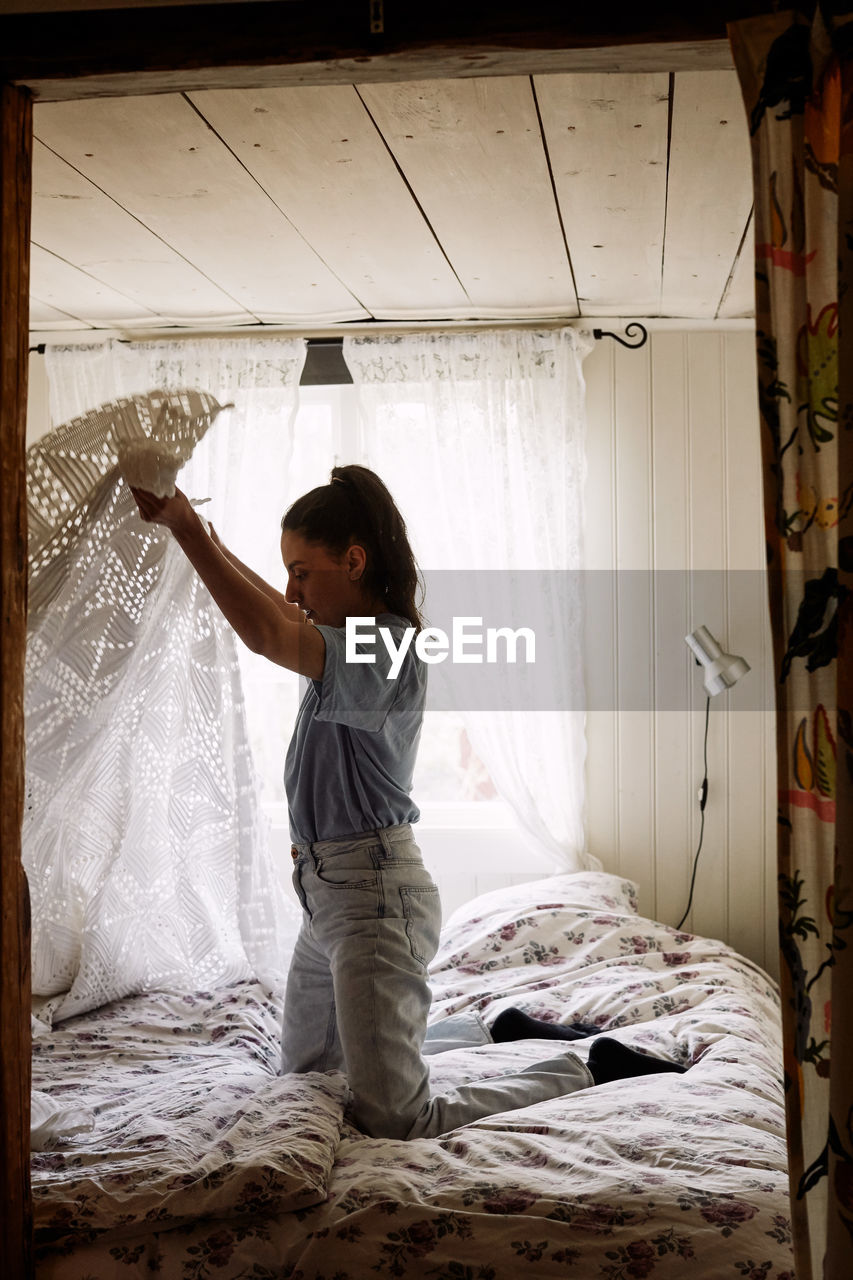 Side view of young woman folding blanket while kneeling on bed in cottage