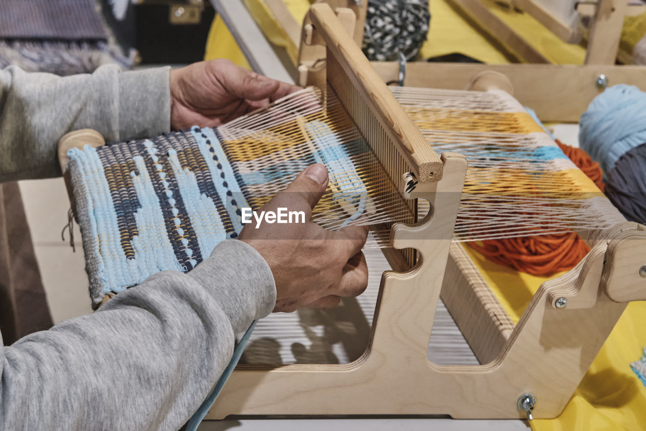 Hands of senior man weaving small rug with pattern on manual table loom.