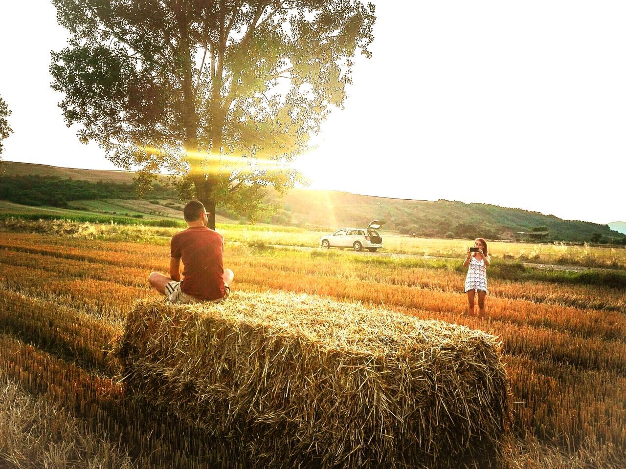 Woman photographing man sitting on hay stack at field against sky during sunset