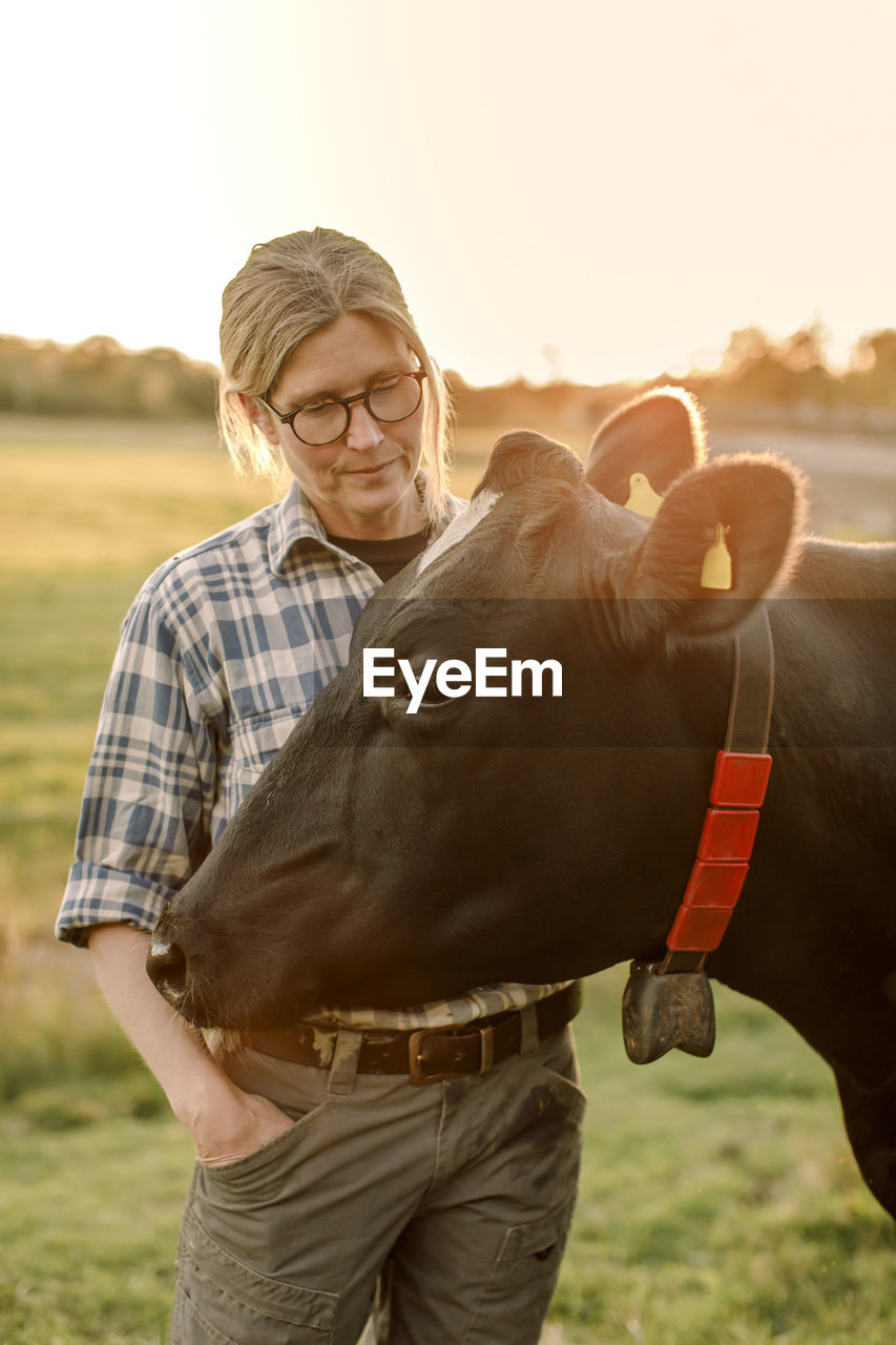 Female farmer standing by cow at field during sunset