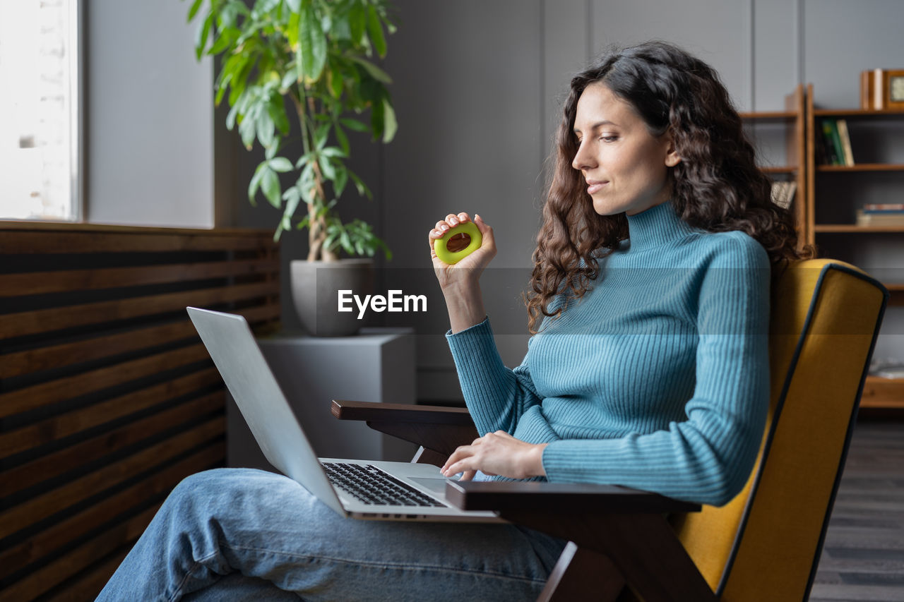 Young satisfied female employee squeezing carpal expander in hand and looking at laptop screen