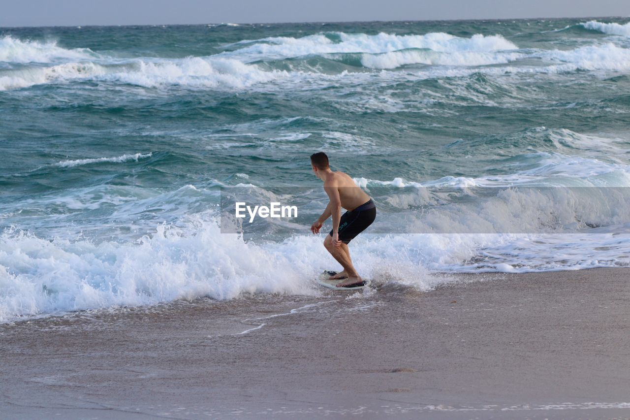 FULL LENGTH OF SHIRTLESS MAN SURFING ON SEA SHORE