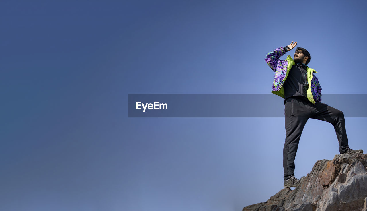 LOW ANGLE VIEW OF MAN STANDING ON ROCK AGAINST CLEAR SKY