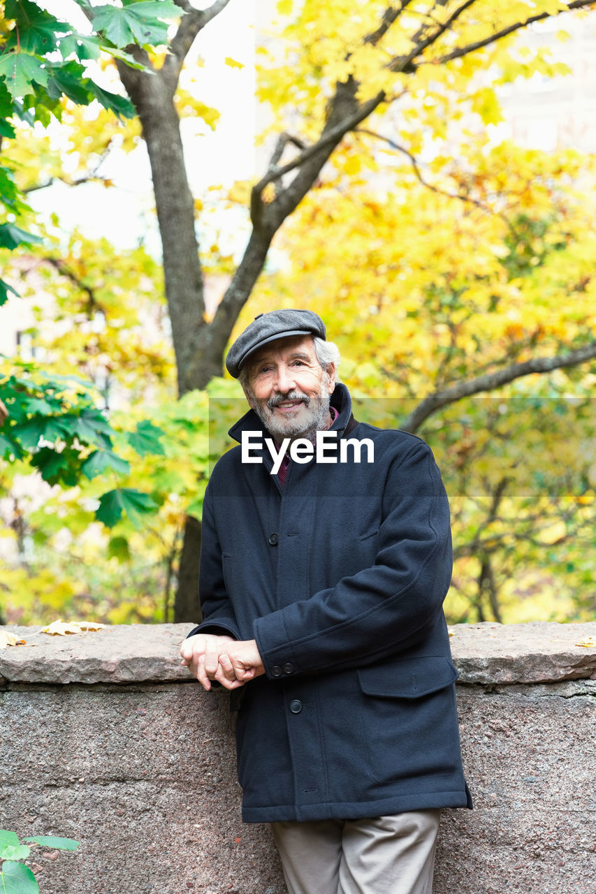 Portrait of confident senior man leaning on retaining wall while standing in park during autumn