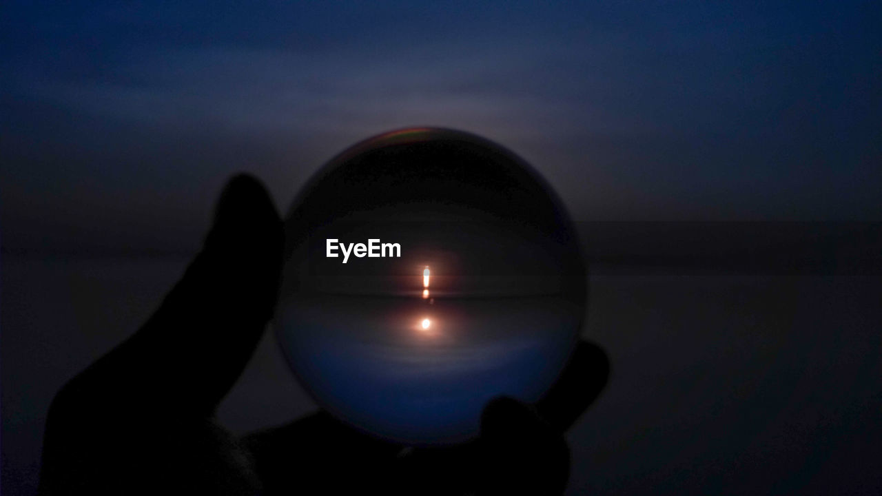 Silhouette hand holding crystal ball with reflection of sea against sky during sunset