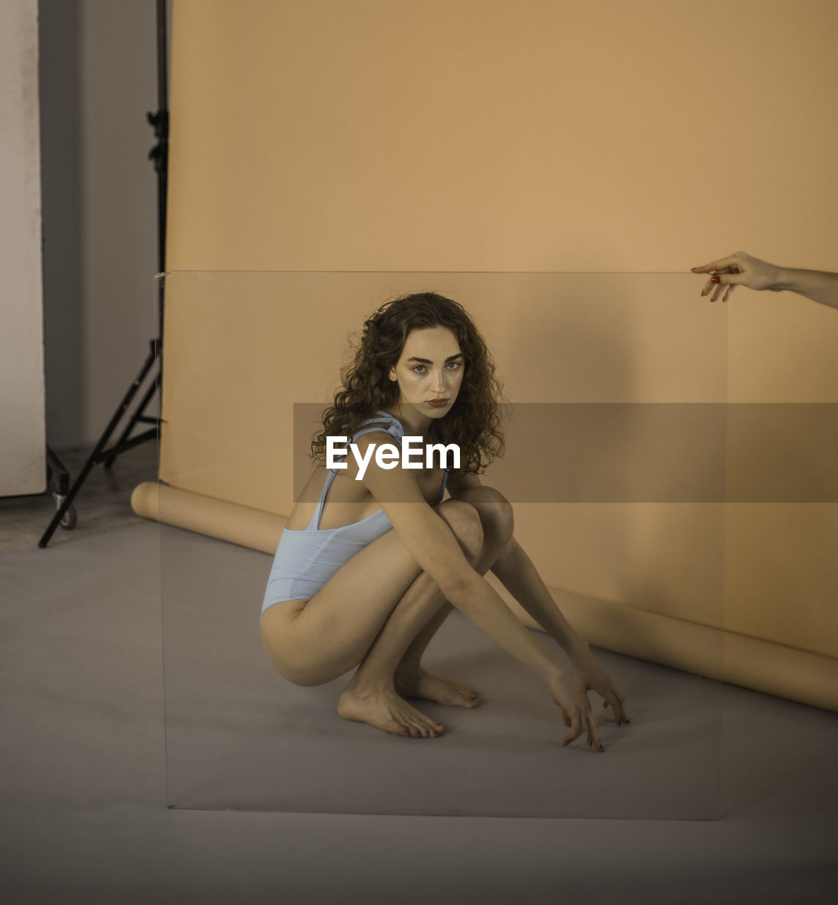 Portrait of young woman crouching seen through glass held by cropped hand during shoot at studio