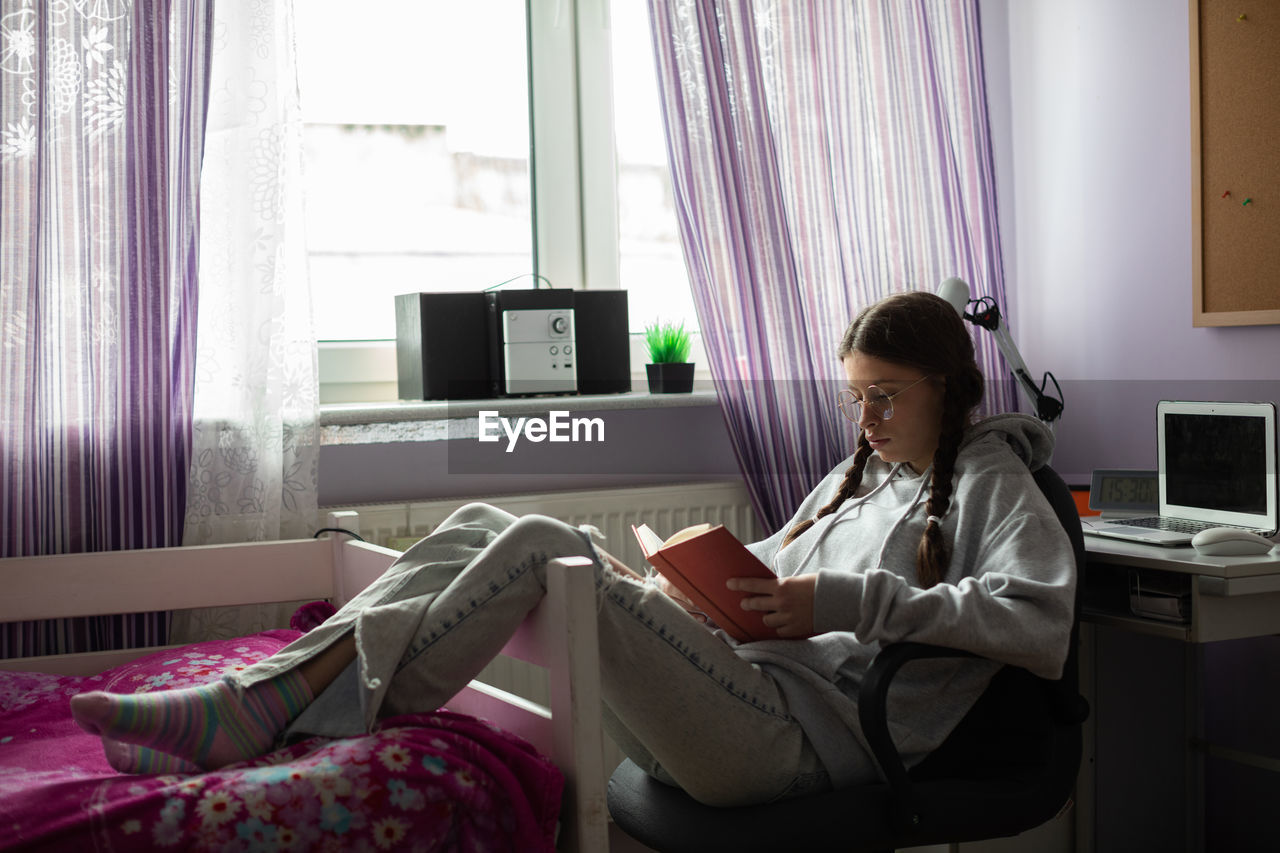 A teenage girl reads her school reading while sitting comfortably by the window in her room.