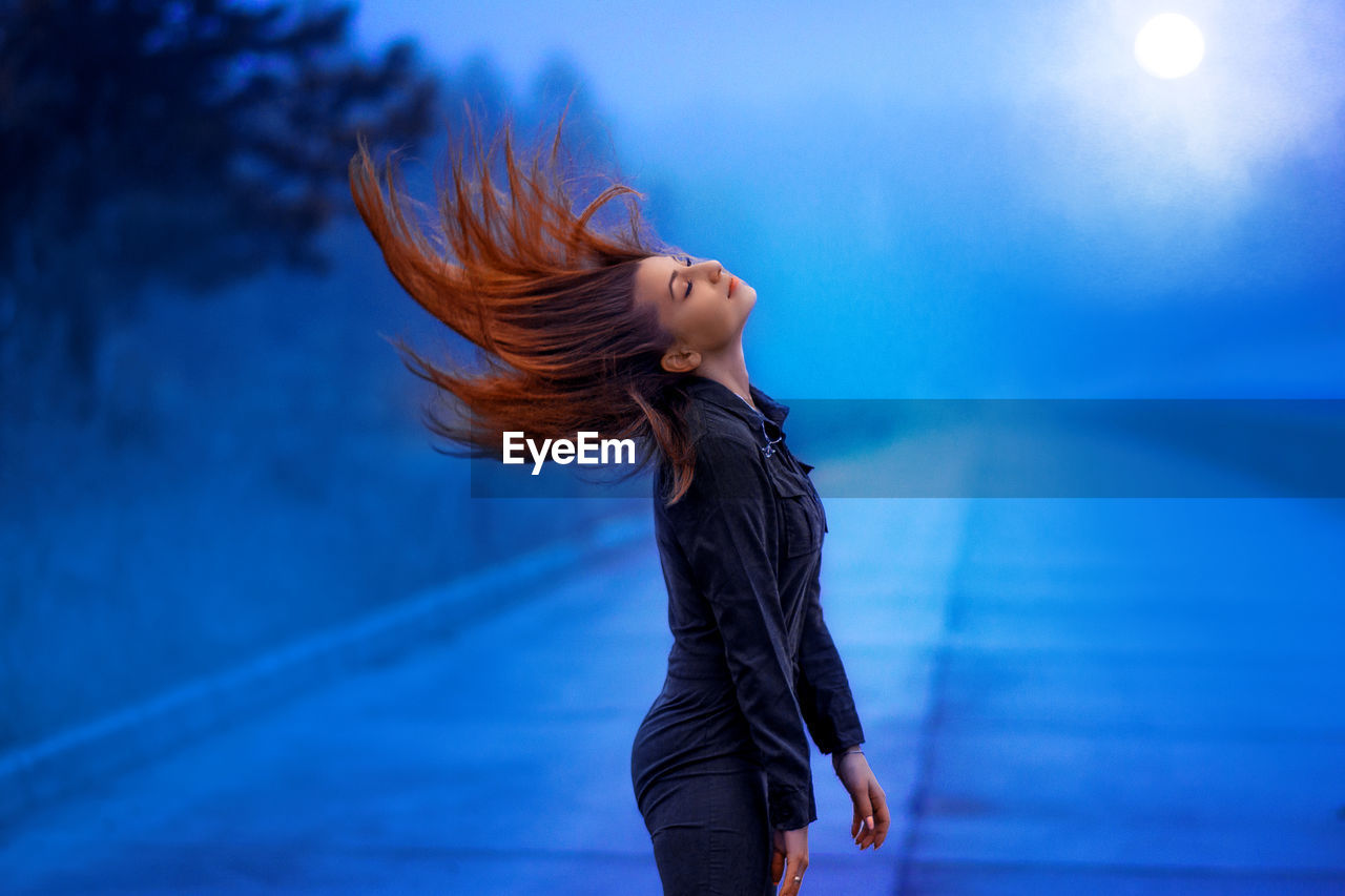 Side view of woman tossing hair while standing on road against sky at dusk