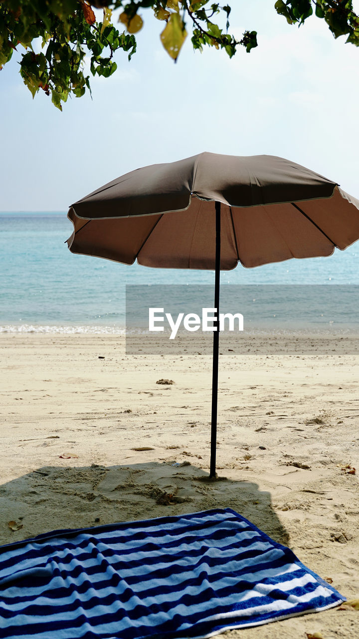 umbrella, beach, land, water, parasol, protection, sea, nature, shade, beach umbrella, sunshade, fashion accessory, sand, security, sky, beauty in nature, tranquility, vacation, trip, relaxation, summer, holiday, tree, tranquil scene, day, horizon over water, scenics - nature, sunlight, horizon, outdoors, no people, chair, blue, travel destinations, plant, idyllic, ocean, travel, lounge chair, absence, seat, shadow