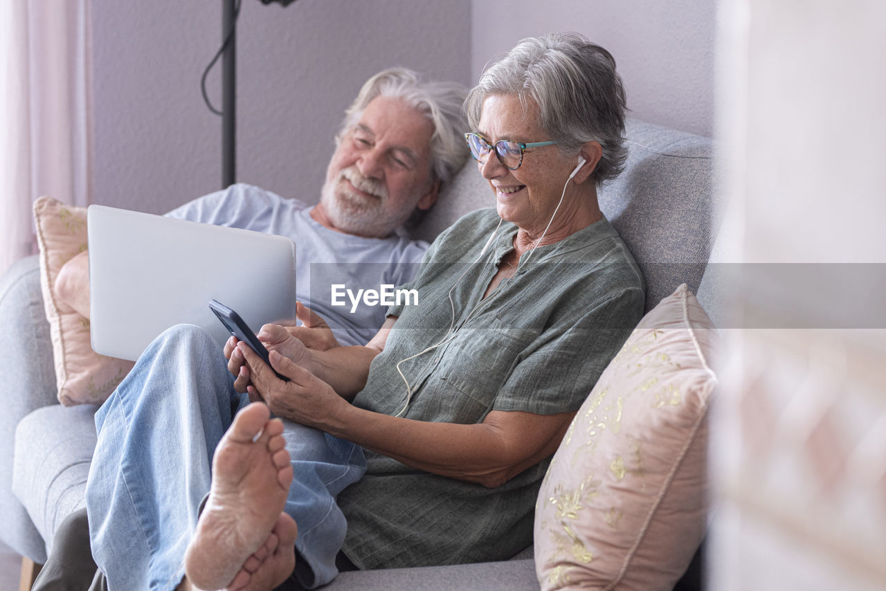 Smiling man using laptop by woman listening music on sofa in living room