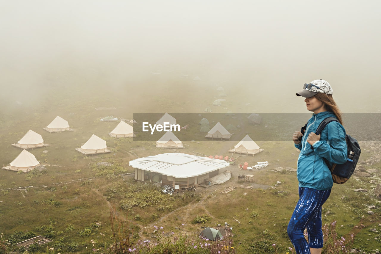 Young woman in cap blue sportswear with backpack hiker  in mountain  against tent camping in fog