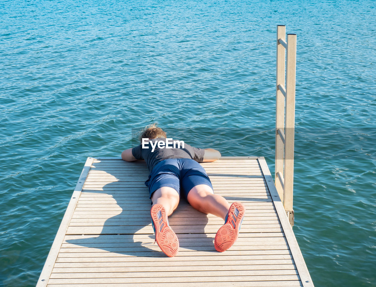 Boy lies on the mole and watches the fish in the lake water below. the joys of childhood