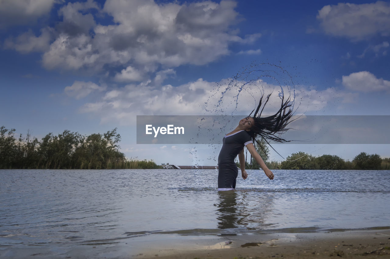 Teenage girl throwing wet hair back in lake against cloudy sky