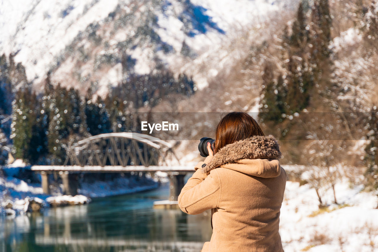 Woman photographing bridge over river against snowcapped mountains