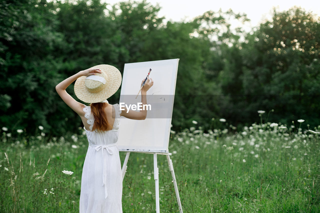 REAR VIEW OF A WOMAN HOLDING HAT ON FIELD