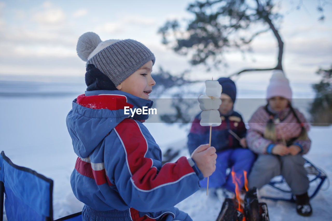 Happy friends sit around campfire on shore of frozen lake in forest and fry marshmallows