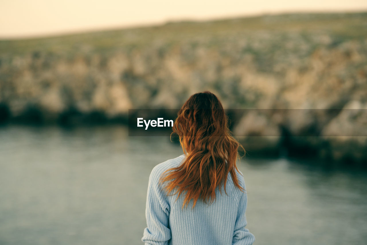 REAR VIEW OF WOMAN STANDING AGAINST SEA DURING SUNSET