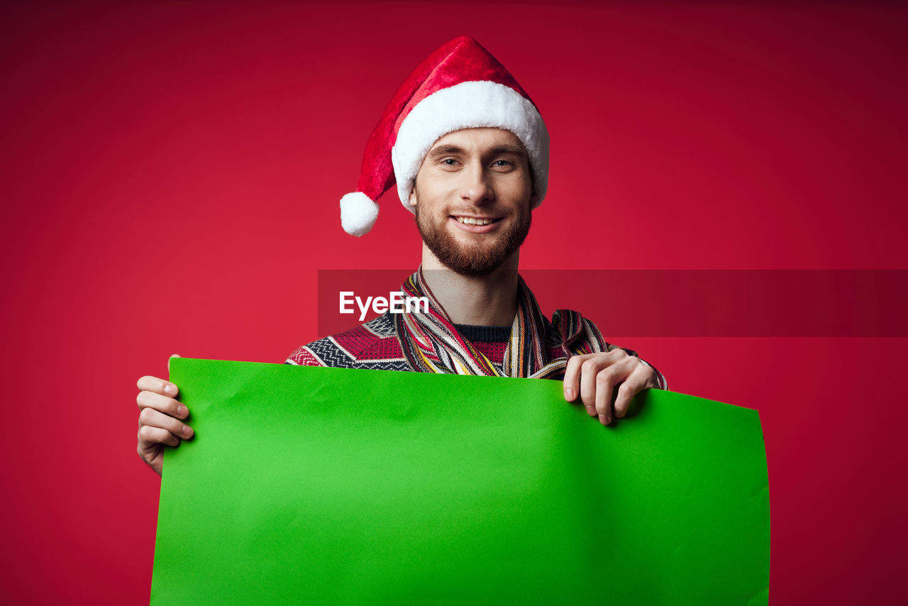 happiness, one person, studio shot, smiling, red, portrait, adult, green, colored background, emotion, beard, celebration, facial hair, indoors, men, cheerful, young adult, christmas, person, clothing, hat, fun, copy space, positive emotion, holiday, looking at camera, santa hat, holding, human face, casual clothing, waist up, gift