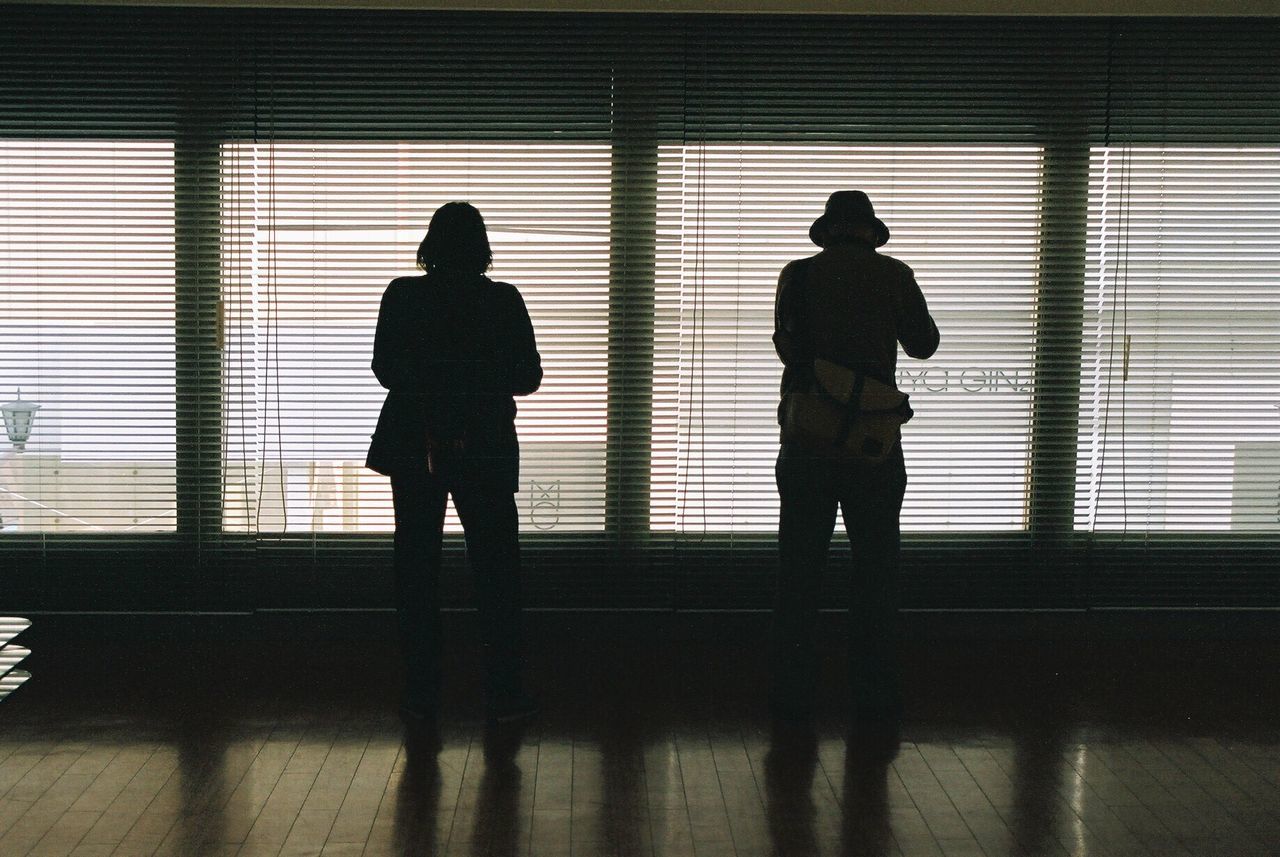 Rear view of two people standing side by side against window