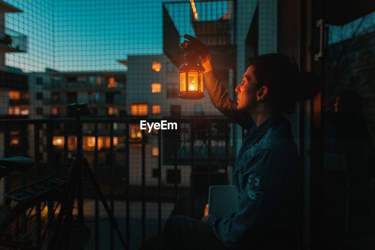 Side view of woman holding oil lamp while sitting in balcony at dusk