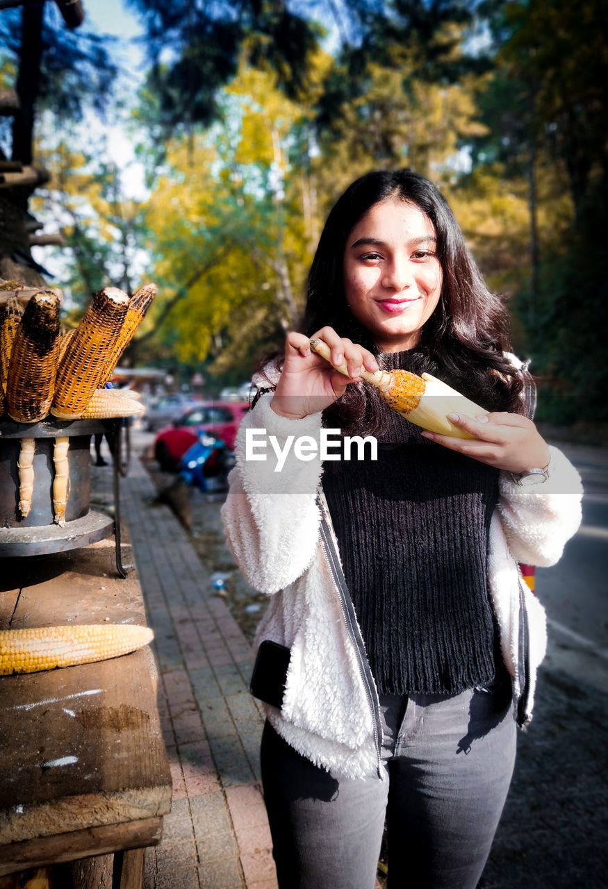 Portrait of smiling young woman having corn at market stall in city