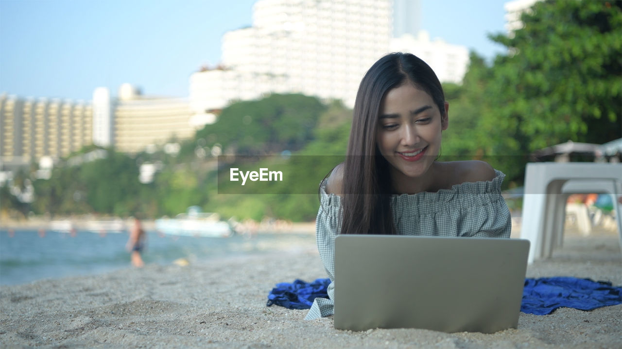 SMILING YOUNG WOMAN USING PHONE WHILE SITTING ON LAPTOP AT PARK