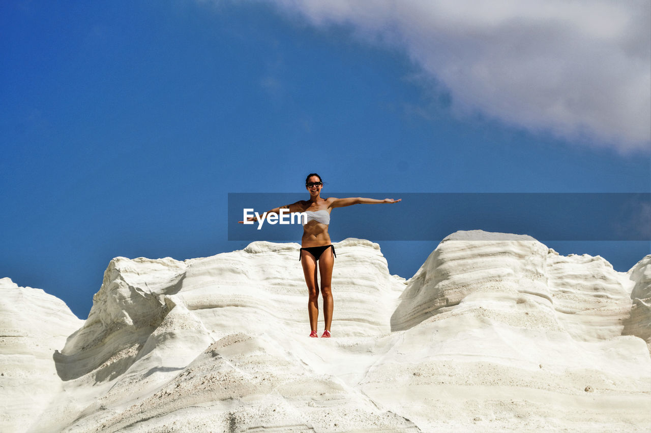 Low angle view of young woman wearing bikini while standing on rock formation against sky during sunny day