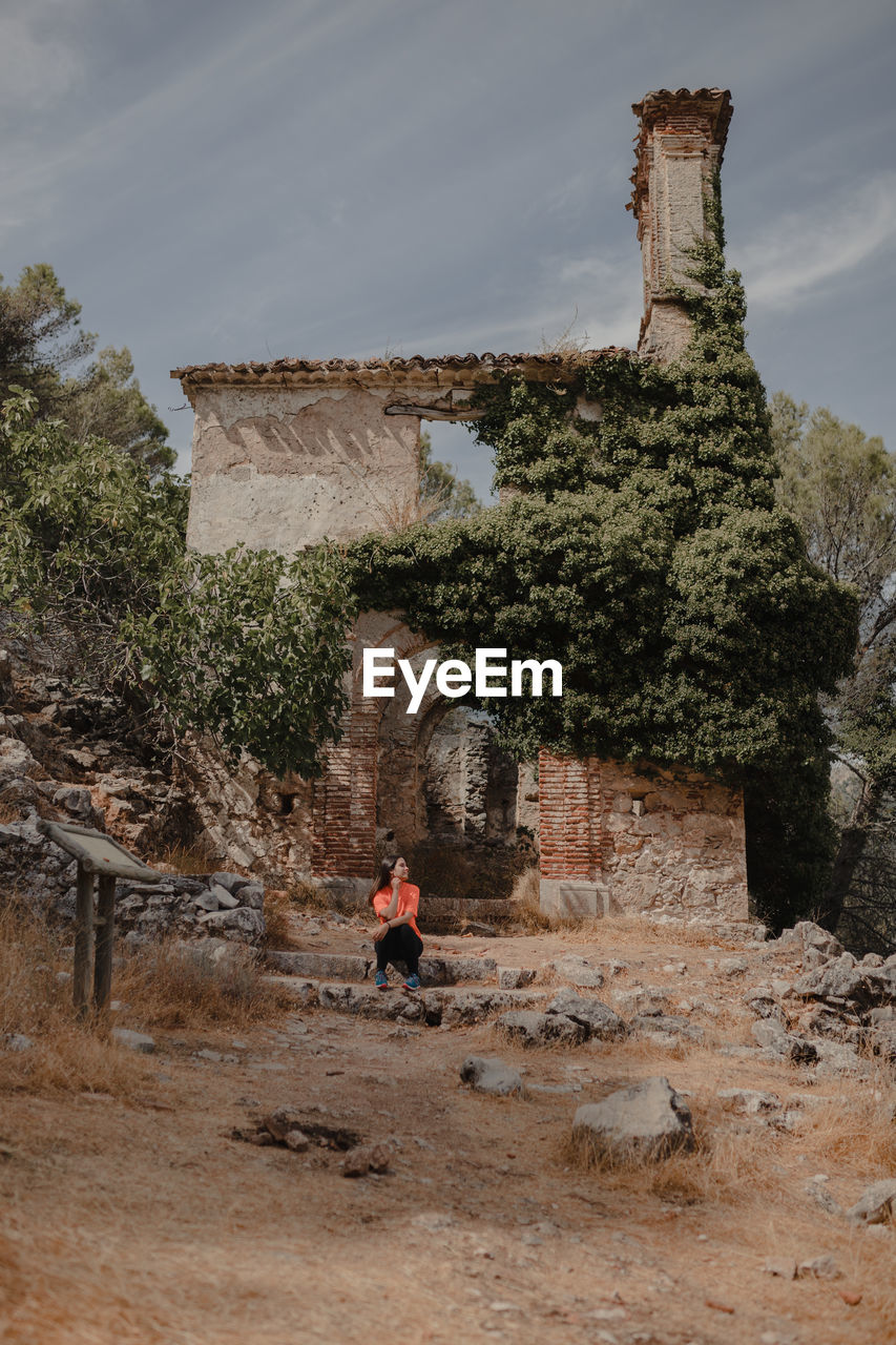 Distant female traveler sitting on stone and observing ancient ruined buildings located in highlands while visiting grazalema village in spain during vacation