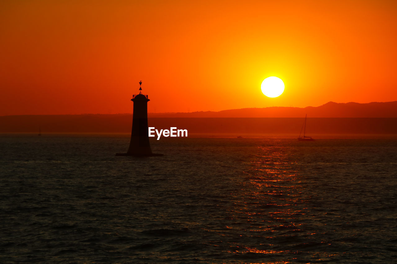 Silhouette lighthouse by sea against orange sky