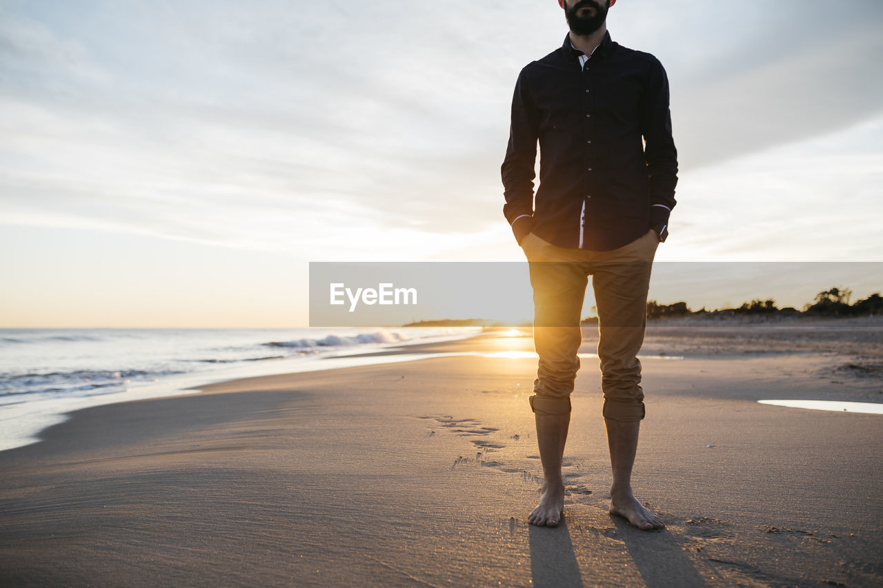 Low section of mid adult man standing at beach against sky during sunset