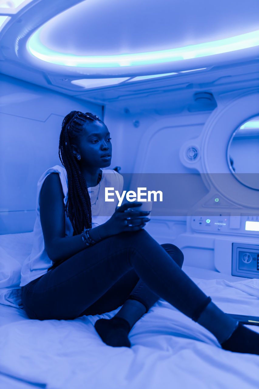 Tranquil black female sitting with cup of drink on bed in room of capsule hotel while resting during vacation