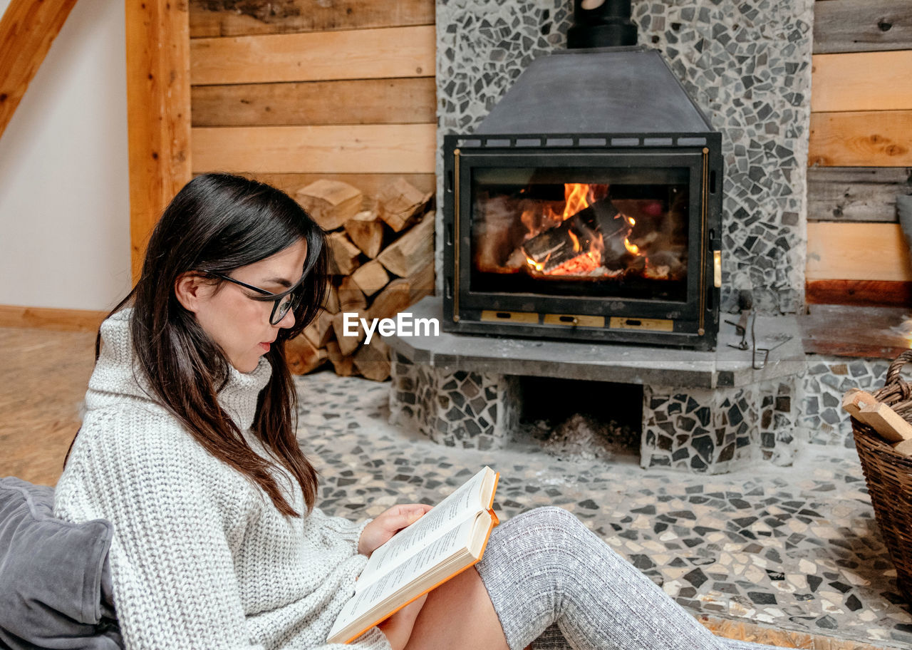 Young woman sitting by fireplace in cozy cabin, reading a book.