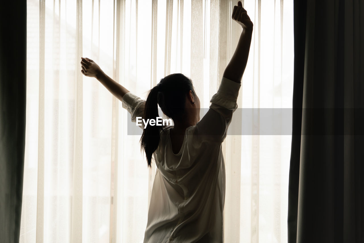 REAR VIEW OF WOMAN WITH ARMS RAISED STANDING IN WINDOW