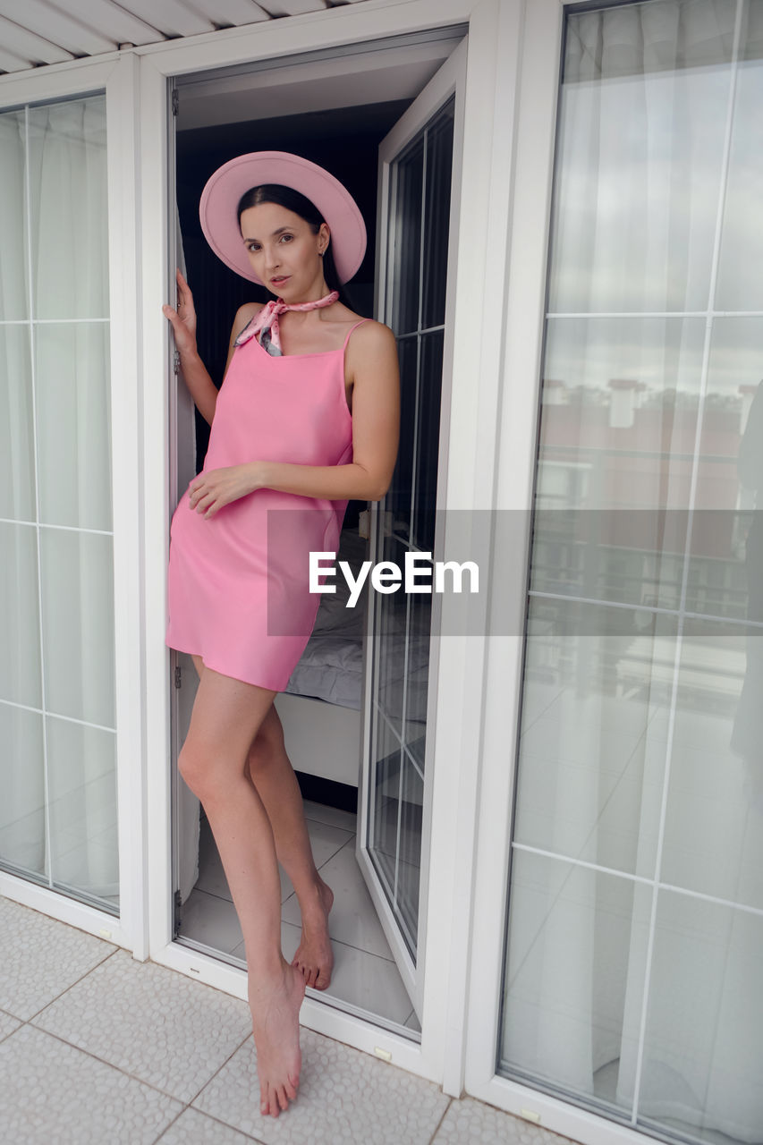 Portrait of a cheerful young woman in a pink dress and hat standing at  door window on the balcony