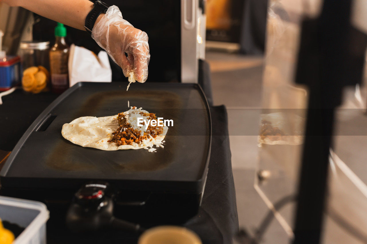 Chef sprinkles a tortilla with cheese preparing fresh quesadilla on black grill.