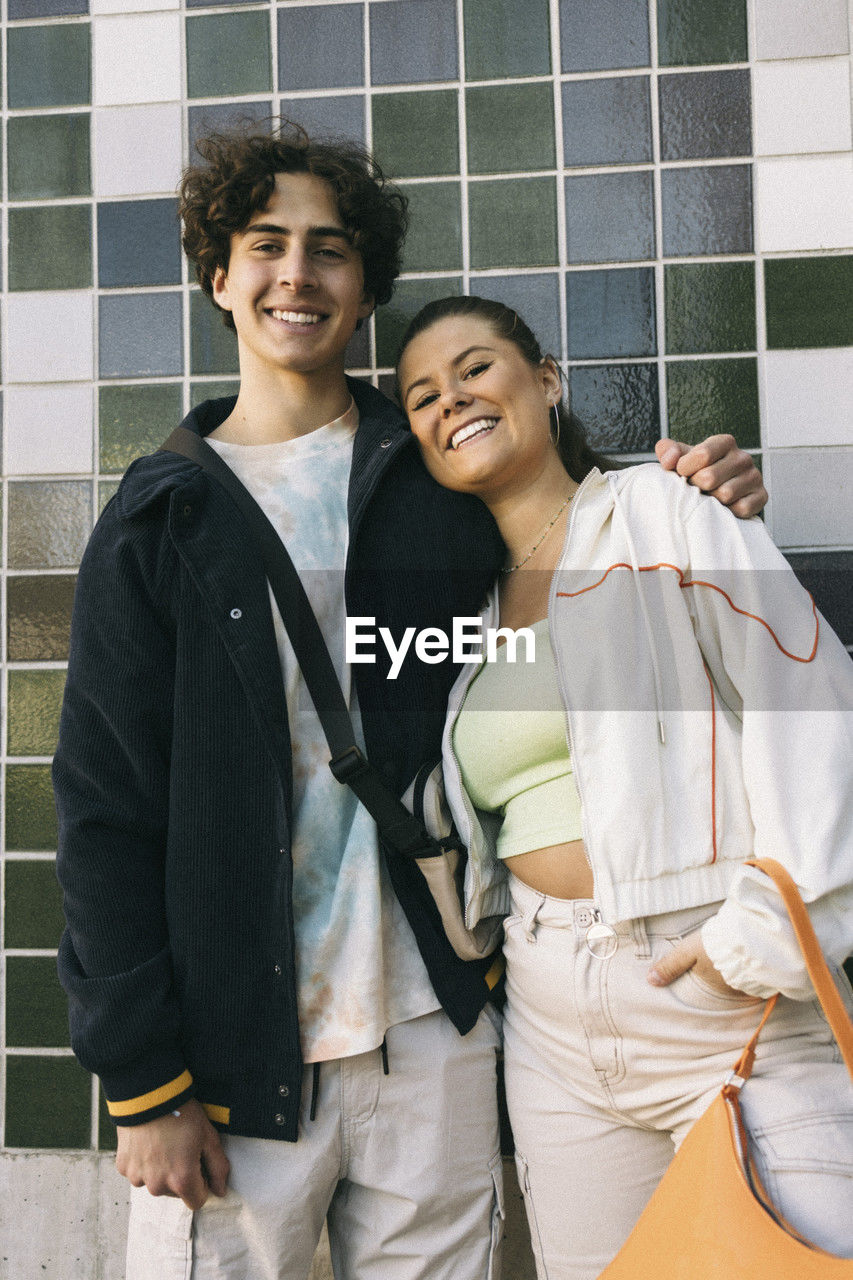 Smiling teenage boy standing with female friend against tiled wall