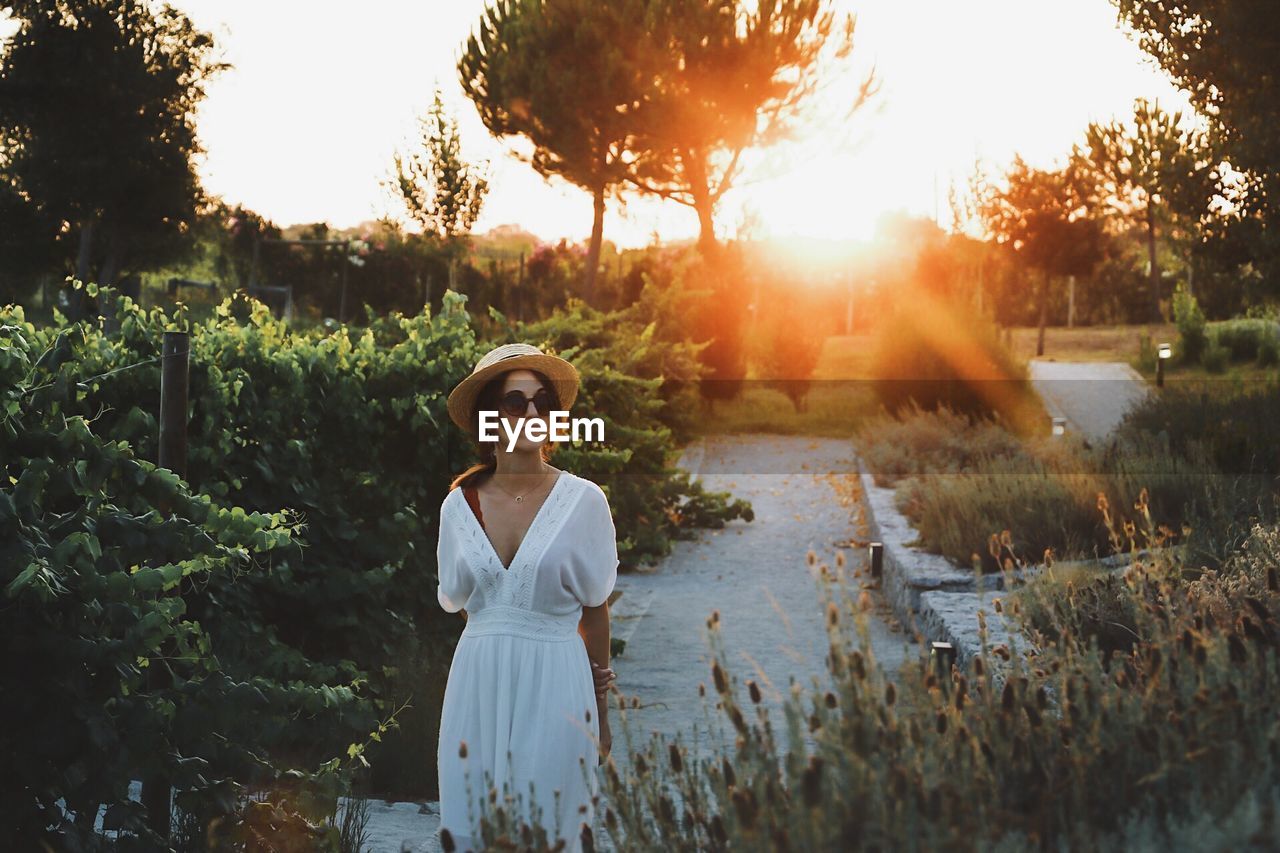 Woman in hat and sunglasses standing against trees during sunset