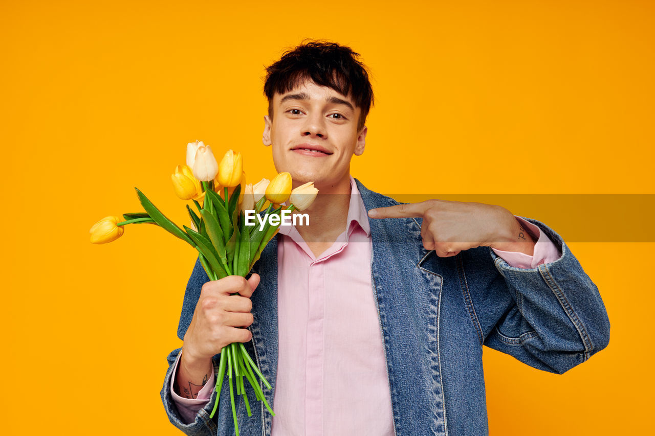 Portrait of young man showing tulip against yellow background