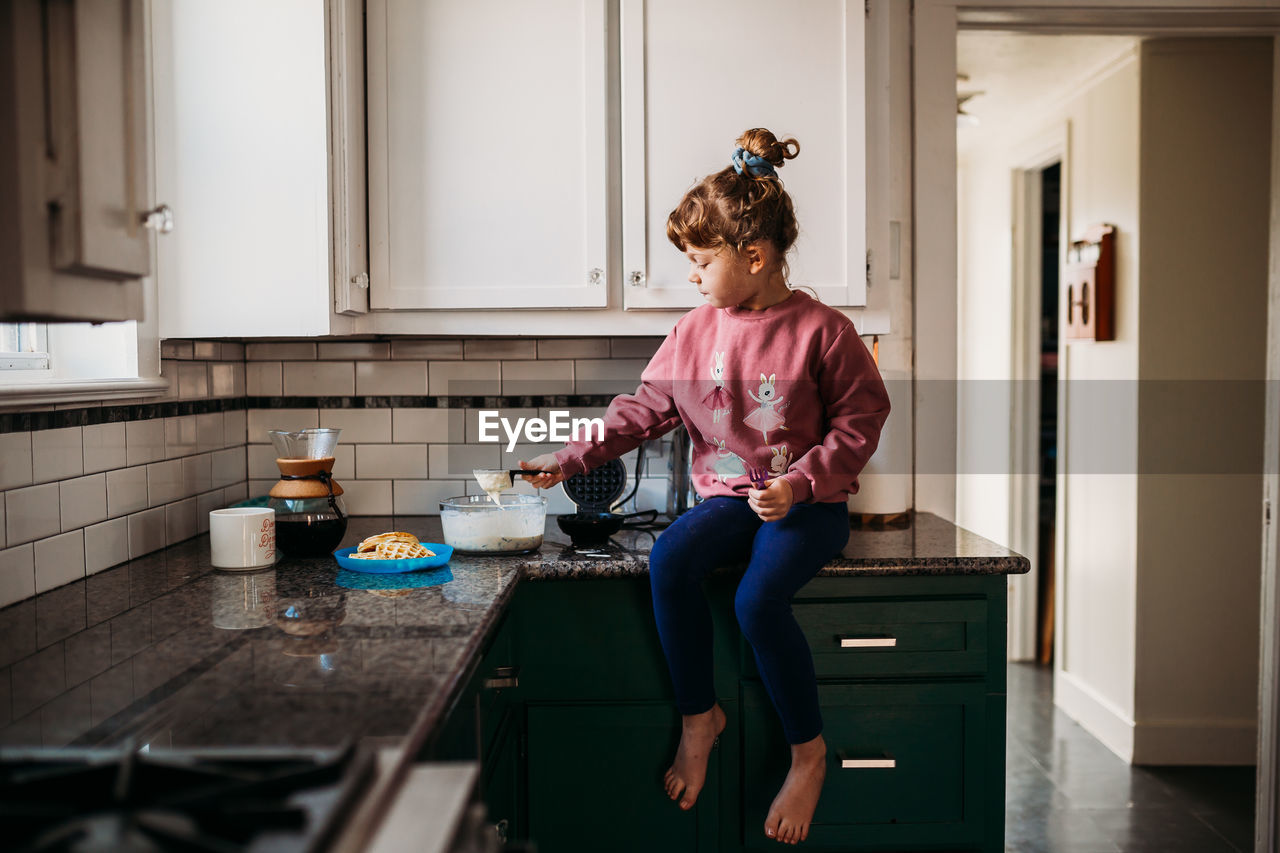 Young girl sitting on kitchen counter scooping waffle batter
